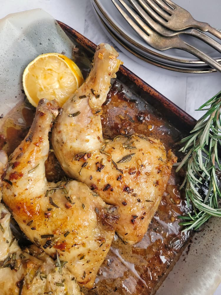 roasted chicken leg quarters on a parchment lined baking sheet with rosemary, lemon and forks and plates in the background