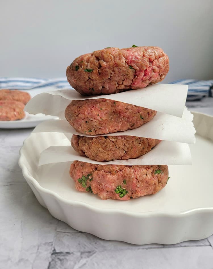 4 raw ground beef patties stacked between parchment paper on a plate, more patties in the background