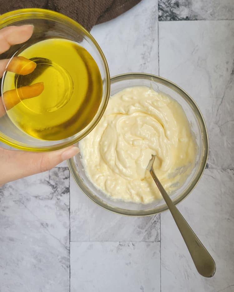 hand holding a ramekin of olive oil over a bowl of aioli with a spoon