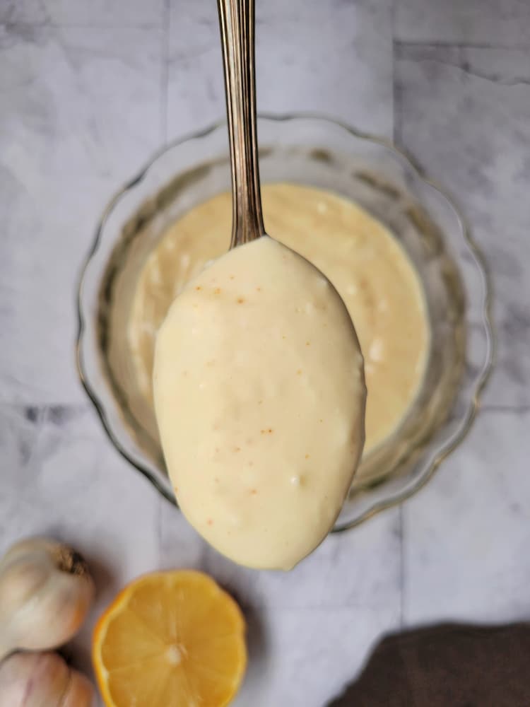 spoon of aioli up in front of the camera over a bowl of it, garlic and lemon in the background
