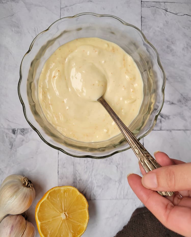 hand with a spoon in a bowl of aioli, lemon and garlic in the background