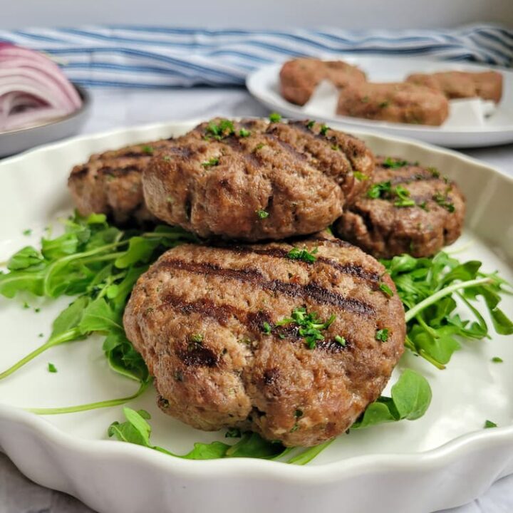 4 cooked grilled beef patties on a bed of arugula on a plate, more patties in the background next to a plate of sliced red onions