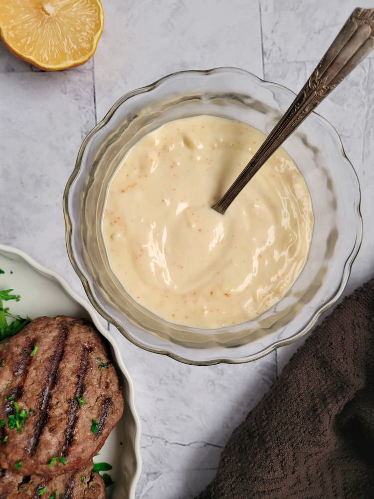 bowl of aioli with a spoon, half a lemon in the background, plate of grilled burgers in the front