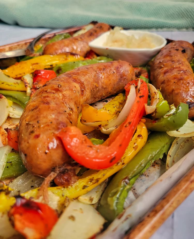 side view of sausage with peppers and onions on a sheet pan, ramekin of sauerkraut in the background