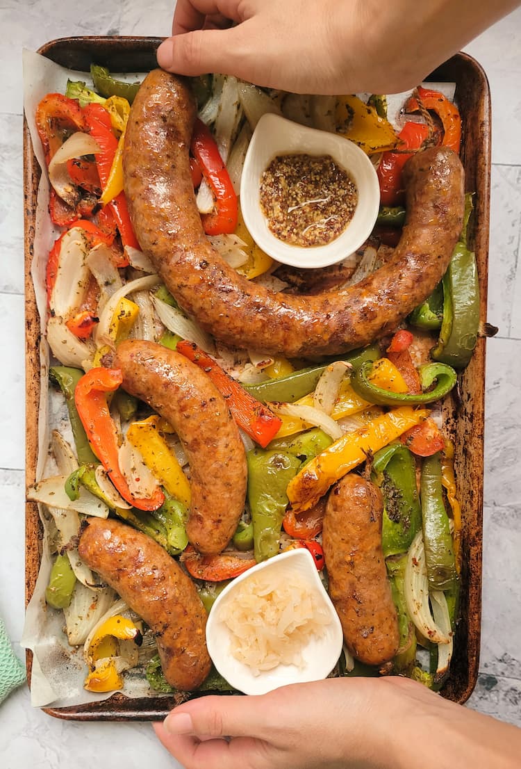 hands holding a sheet pan of sausage with pepper and onions, ramekins of sauerkraut and grainy mustard on the pan
