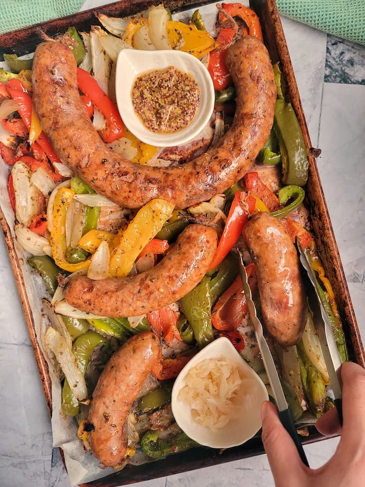 hand with tongs grabbing a sausage of a sheet pan with more over top some cooked peppers and onions, ramekins of sauerkraut and grainy mustard