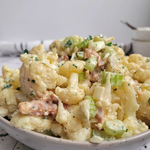 cauliflower 'potato' salad in a bowl with a creamy sauce, celery pieces, bacon, onions and fresh chopped parsley