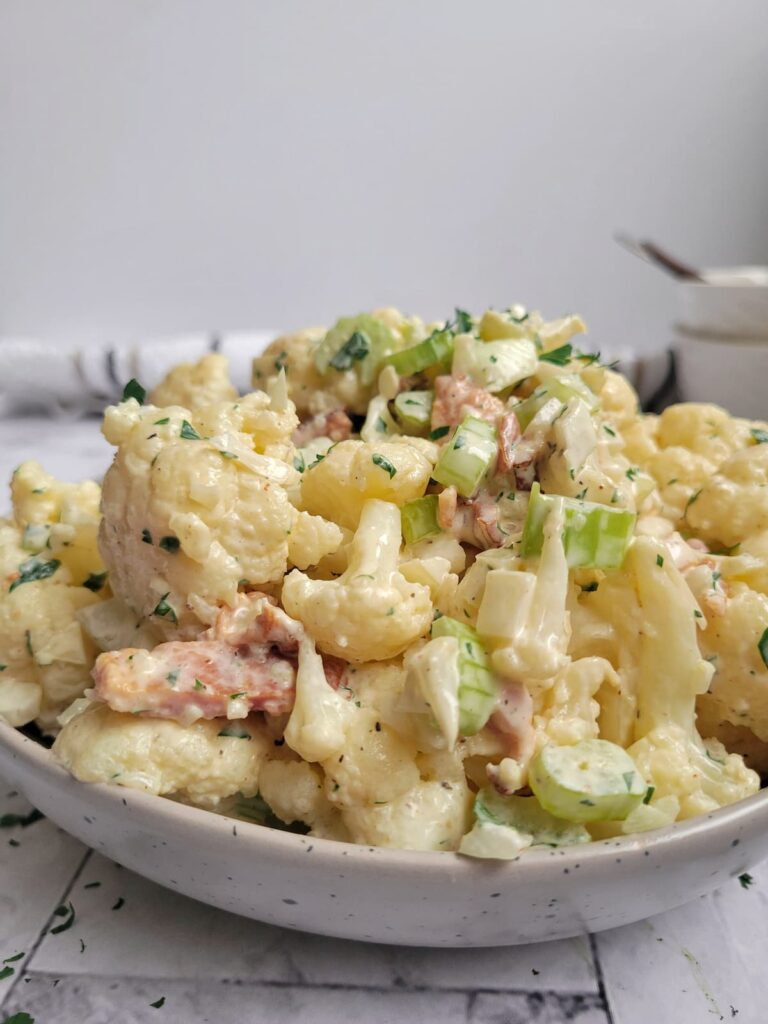 cauliflower 'potato' salad in a bowl with a creamy sauce, celery pieces, bacon, onions and fresh chopped parsley