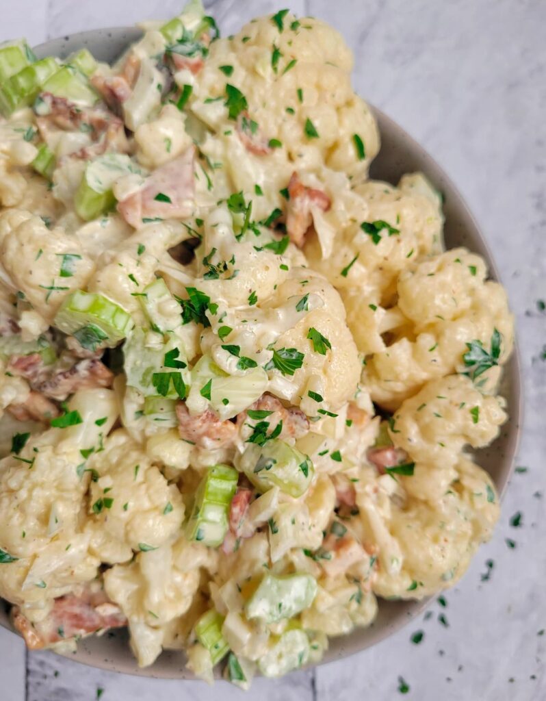cauliflower 'potato' salad in a bowl with a creamy sauce, celery pieces, bacon, onions and fresh chopped parsley