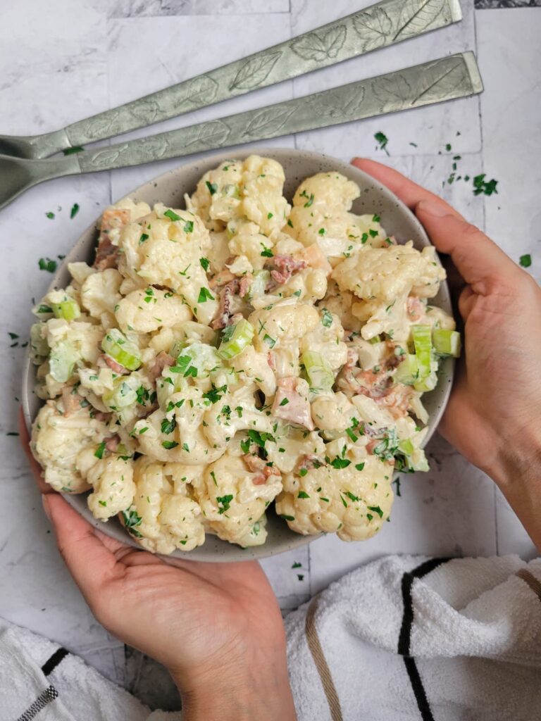 hands holding a bowl of cauliflower 'potato' salad with bacon, celery and fresh chopped parsley, handles of 2 big spoons in the background