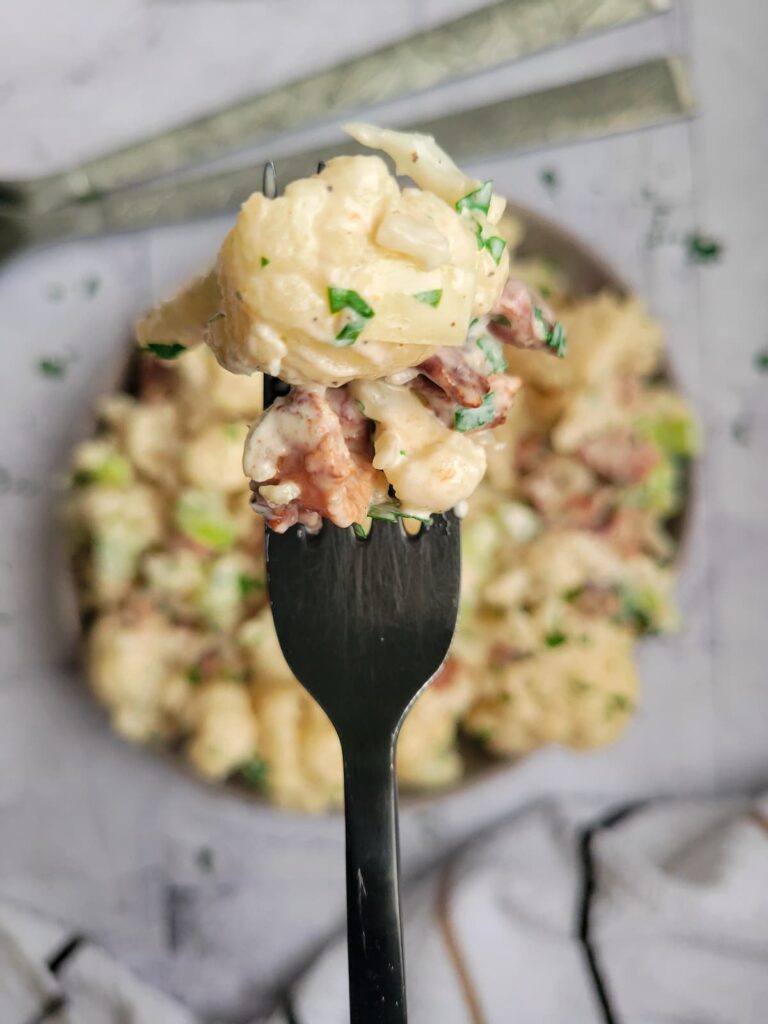 cauliflower floret in a creamy sauce on a fork with some fresh chopped parsley and bacon over a bowl of the rest