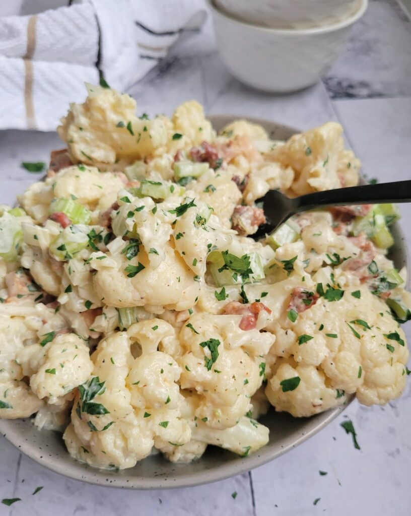 cauliflower 'potato' salad in a bowl with a creamy sauce, celery pieces, bacon, onions and fresh chopped parsley, fork in the bowl, two stacked empty bowls in the background