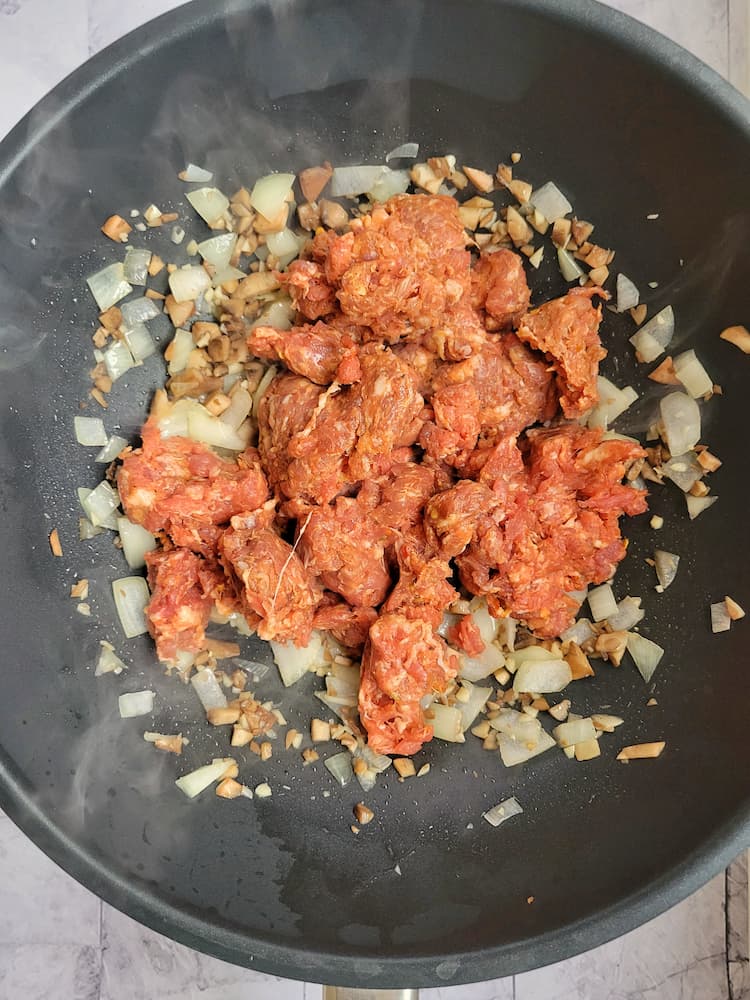 raw italian sausage in a skillet with diced onion, garlic and mushroom stems