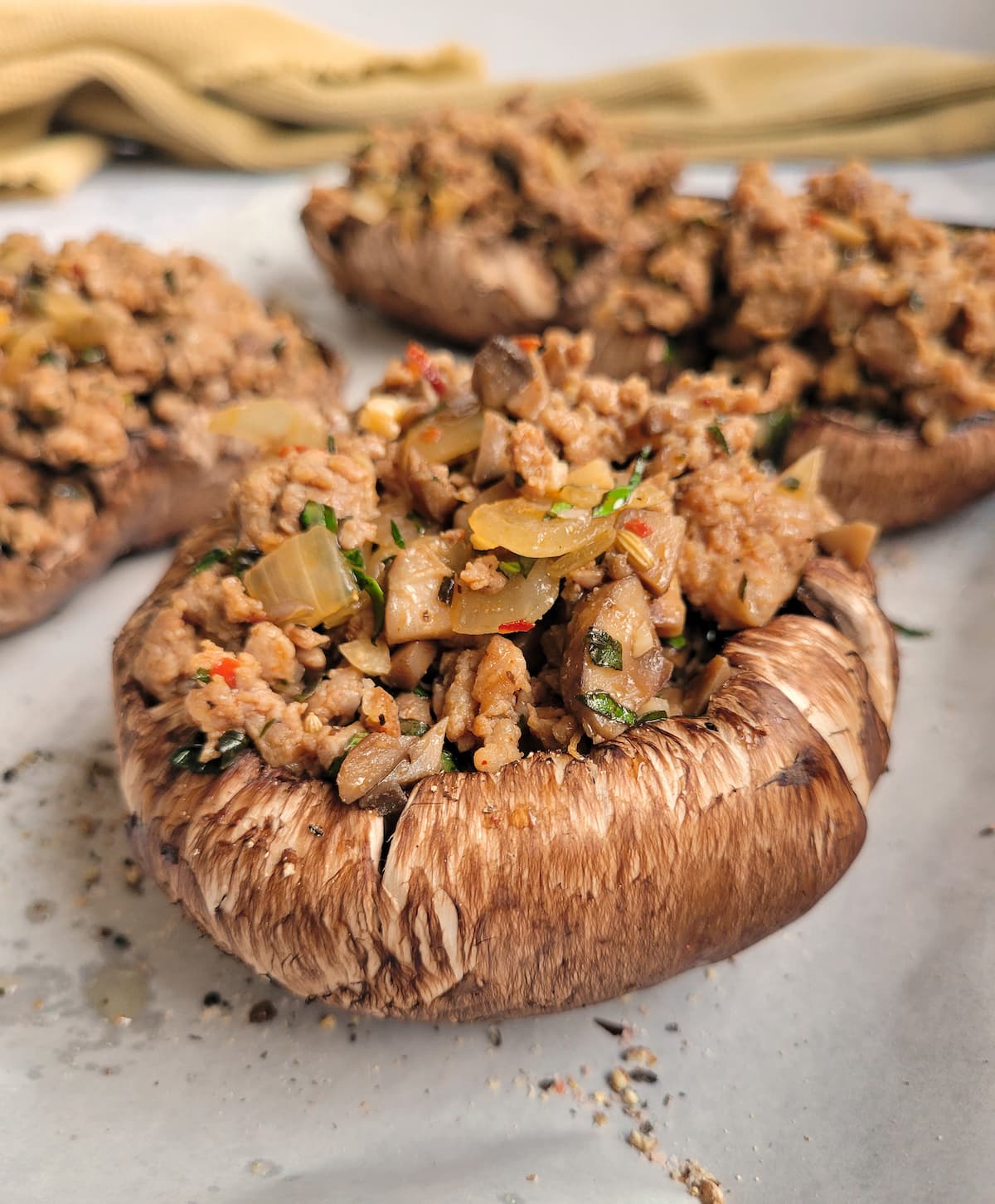 close up of a raw portobello mushroom stuffed with cooked spiced sausage and white onion, more stuffed mushrooms in the background
