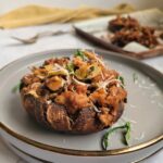 cooked sausage stuffed portobello mushroom on a double plate garnished with fresh parmesan cheese and fresh basil, baking tray with more mushrooms in the background next to some forks.