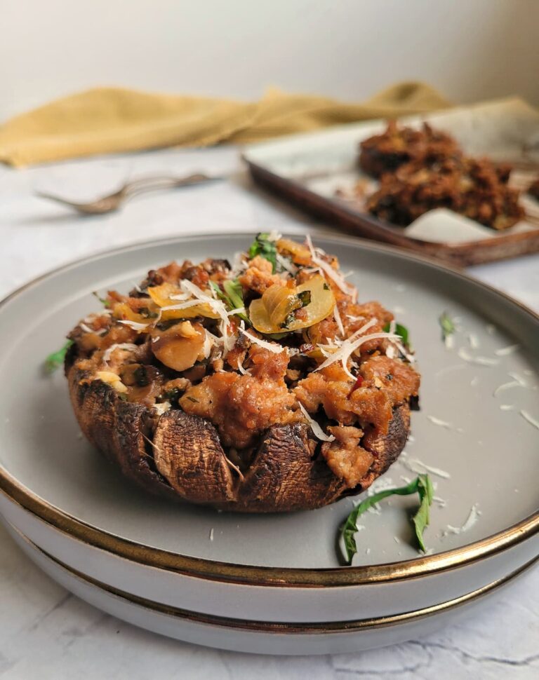 cooked sausage stuffed portobello mushroom on a double plate garnished with fresh parmesan cheese and fresh basil, baking tray with more mushrooms in the background next to some forks.