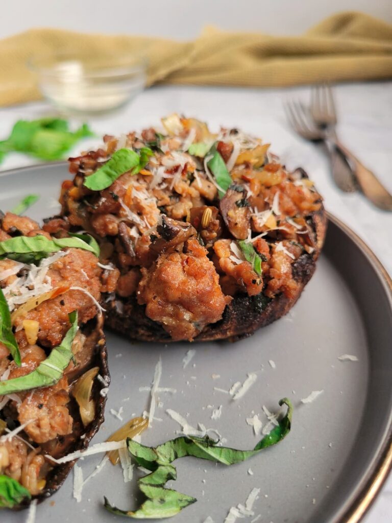 two sausage stuffed portobello mushrooms on a plate, garnished with freshly grated parmesan cheese and fresh basil, two forks in the background