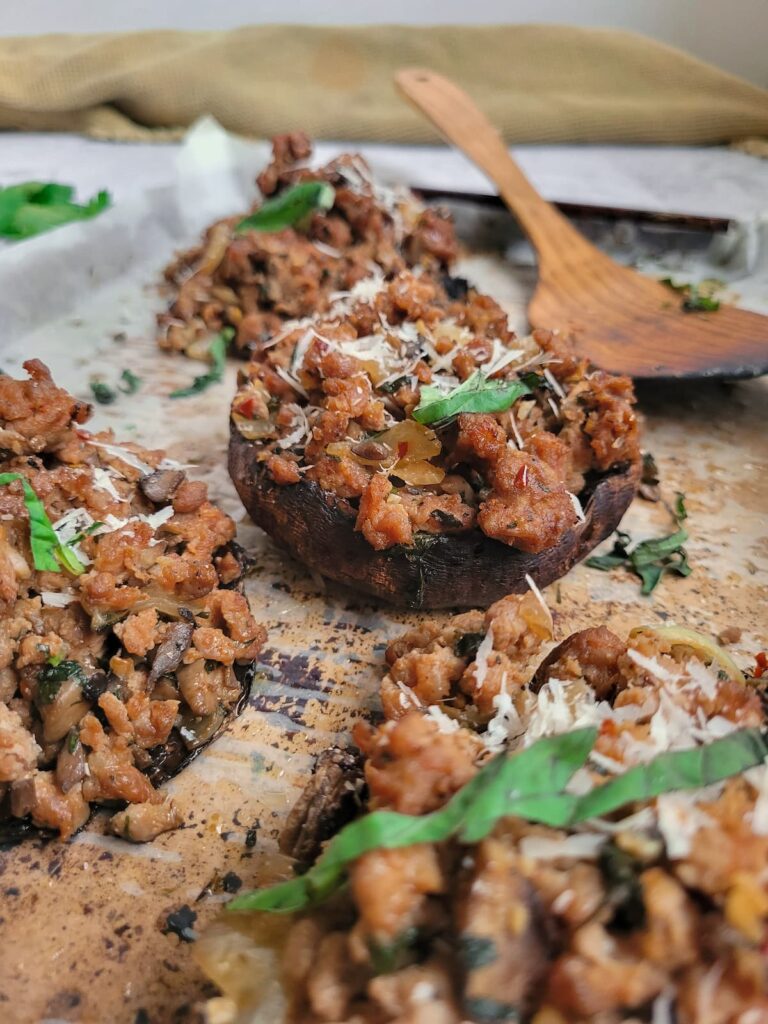cooked stuffed portobello mushrooms with sausage on a lined baking sheet, garnished with fresh basil and parmesan cheese, wooden spoon in the background