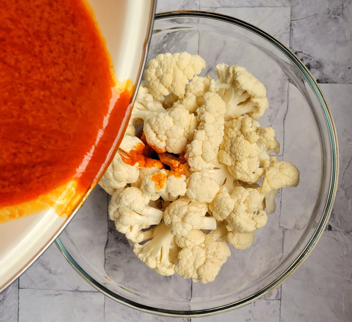 bowl of raw cauliflower florets with buffalo sauce being poured from a pan over top