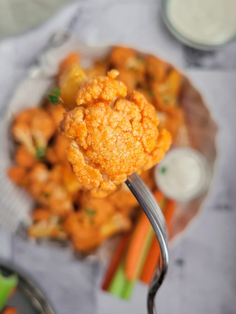 buffalo cauliflower bite on a fork over a plate with the rest of them, blue cheese, celery and carrots in the background