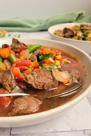 bowl of beef and vegetable soup with beef chunks, celery, tomatoes, beans, carrots and broth, spoon in the bowl, another bowl of soup in the background