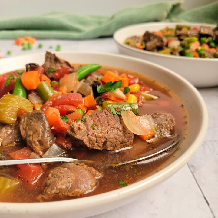 bowl of beef and vegetable soup with beef chunks, celery, tomatoes, beans, carrots and broth, spoon in the bowl, another bowl of soup in the background