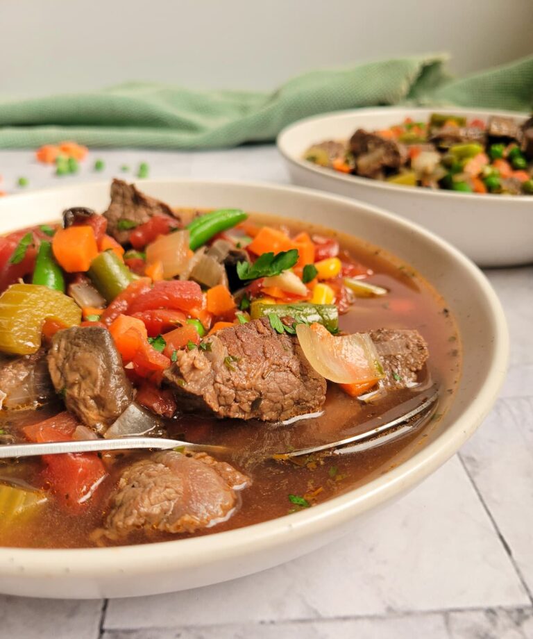 bowl of beef and vegetable soup with beef chunks, celery, tomatoes, beans, carrots and broth, spoon in the bowl, another bowl of soup in the background