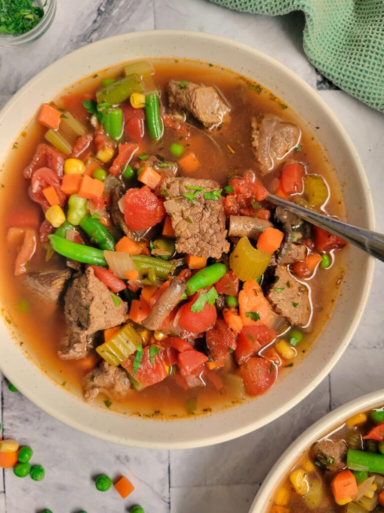 bowl of beef and vegetable soup with beef chunks, celery, tomatoes, beans, carrots and broth, spoon in the bowl, another bowl in the bottom corner