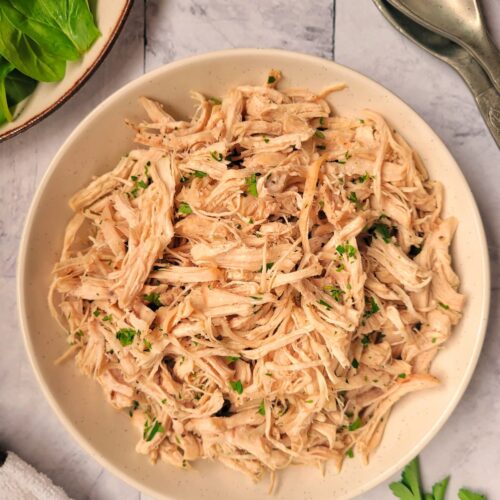 bowl of shredded chicken garnished with fresh chopped parsley, greens and serving spoons in the background