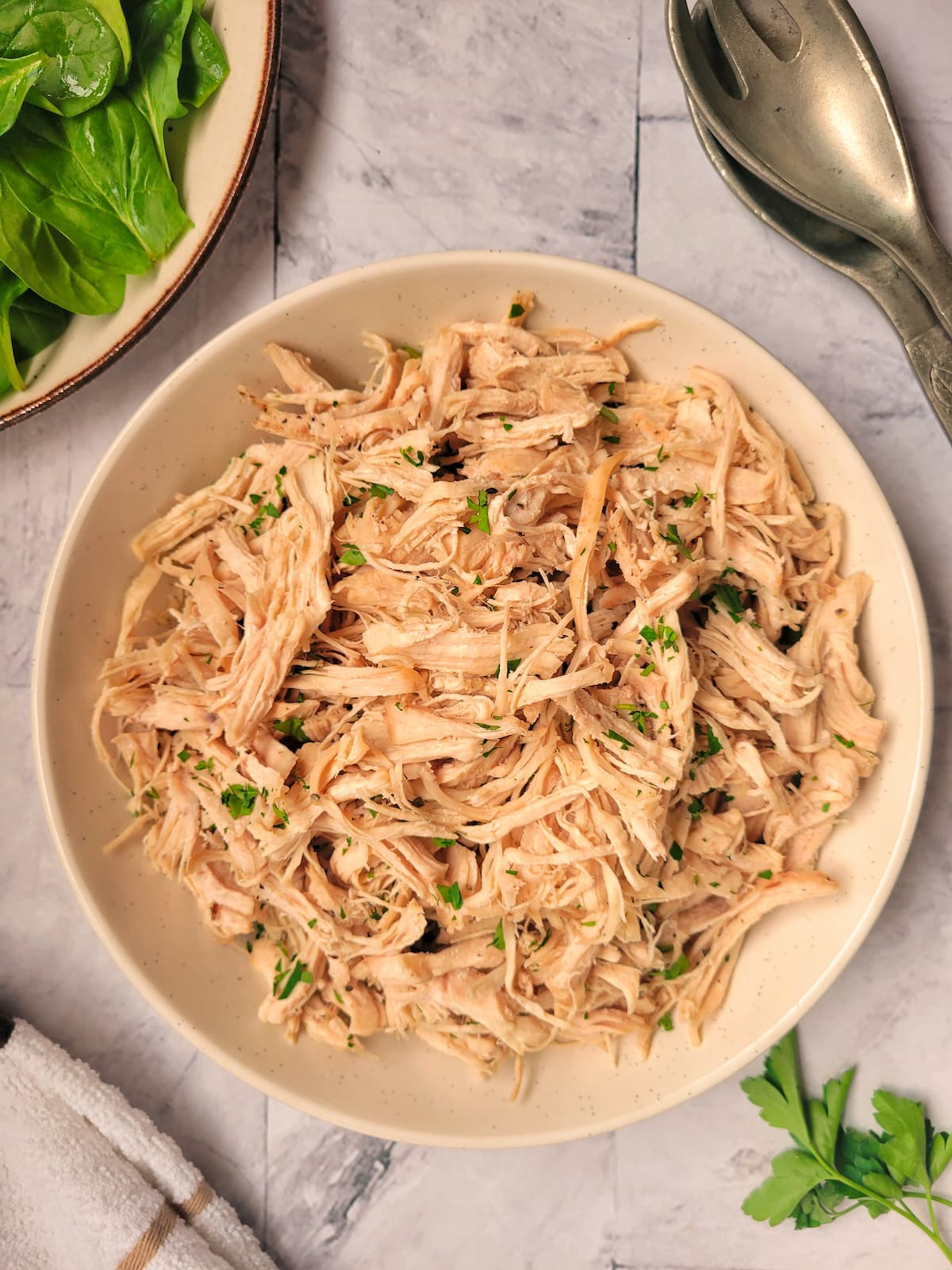 bowl of shredded chicken garnished with fresh chopped parsley, greens and serving spoons in the background