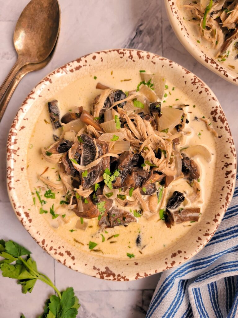 bowl of creamy mushrooms soup with shredded chicken, garnished with fresh parsley, another bowl of soup in the background, spoons on the side