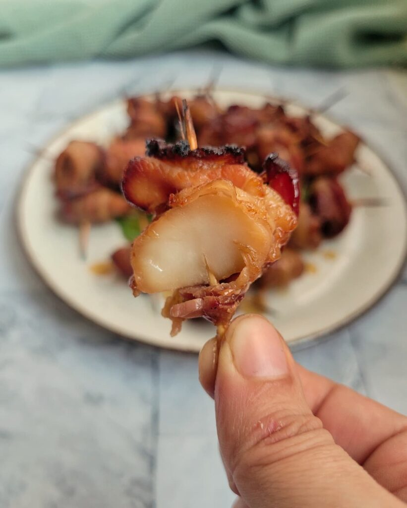 hand holding a half bitten bacon wrapped water chestnut with a toothpick in front of a plate with the rest