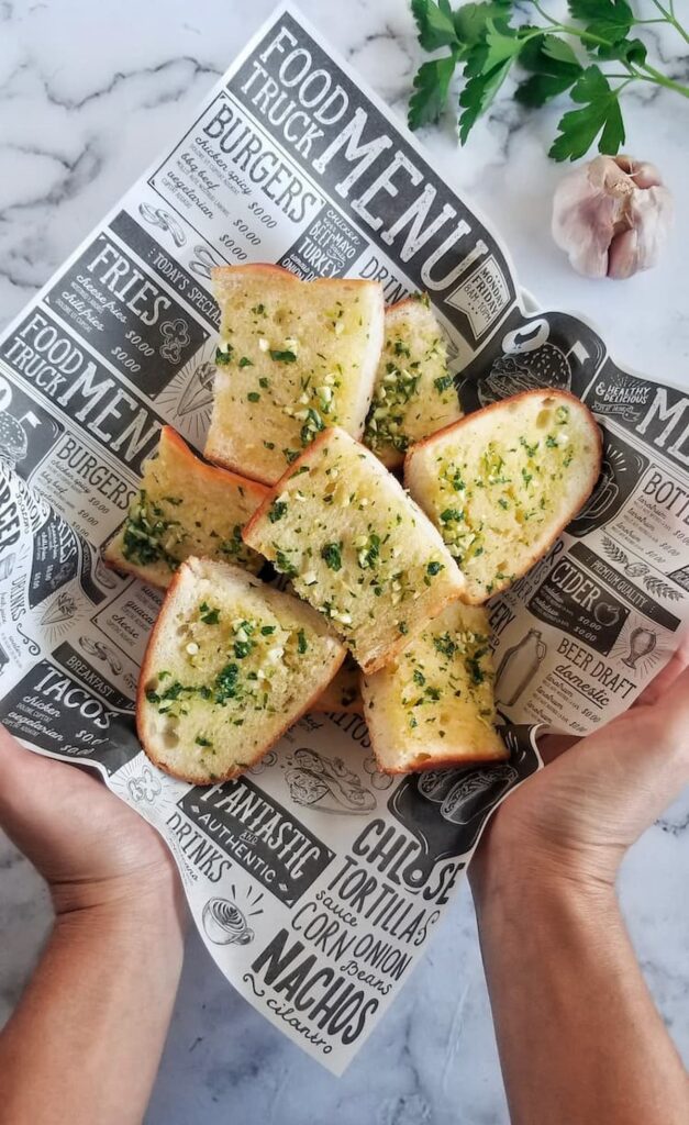 hands holding a basket of fresh sliced garlic bread with fresh minced garlic and chopped parsley, bulb of garlic and fresh parsley in the background