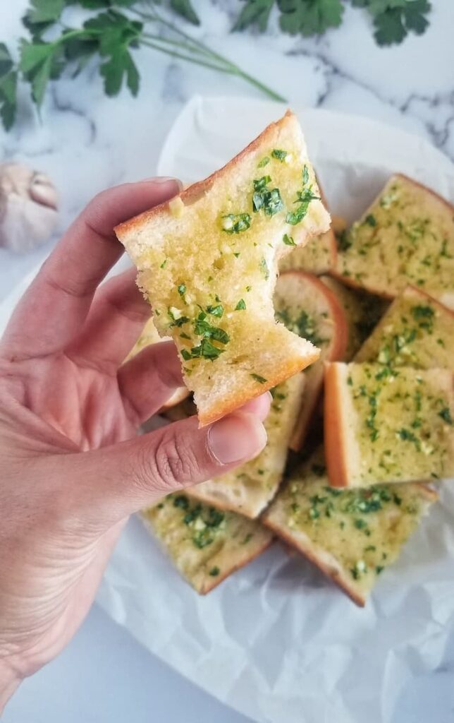 hand holding up a bitten piece of garlic bread with fresh parsley and minced garlic