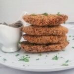 four chickpea patties stacked on top of each other on a plate next to a white gravy boat holding vegan mushroom gravy, garnished with fresh chopped parsley