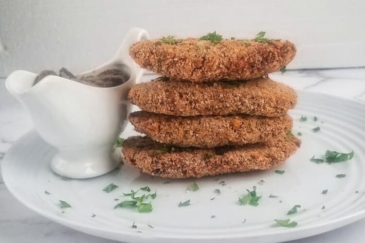 four chickpea patties stacked on top of each other on a plate next to a white gravy boat holding vegan mushroom gravy, garnished with fresh chopped parsley