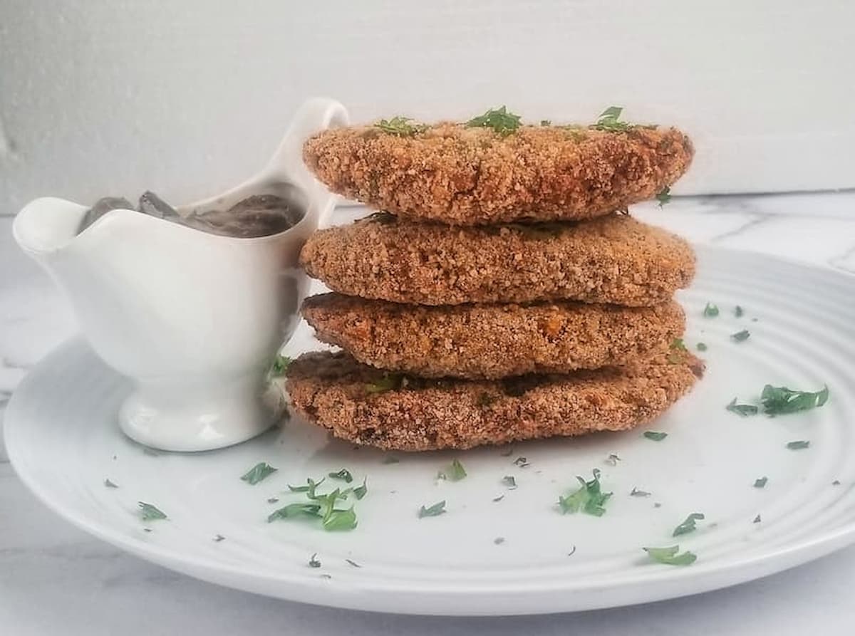 four chickpea patties stacked on top of each other on a plate next to a white gravy boat holding vegan mushroom gravy, garnished with fresh chopped parsley