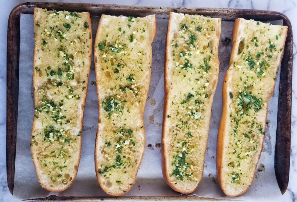 parchment lined baking sheet with four loaves of fresh garlic bread with fresh chopped parsley and minced garlic