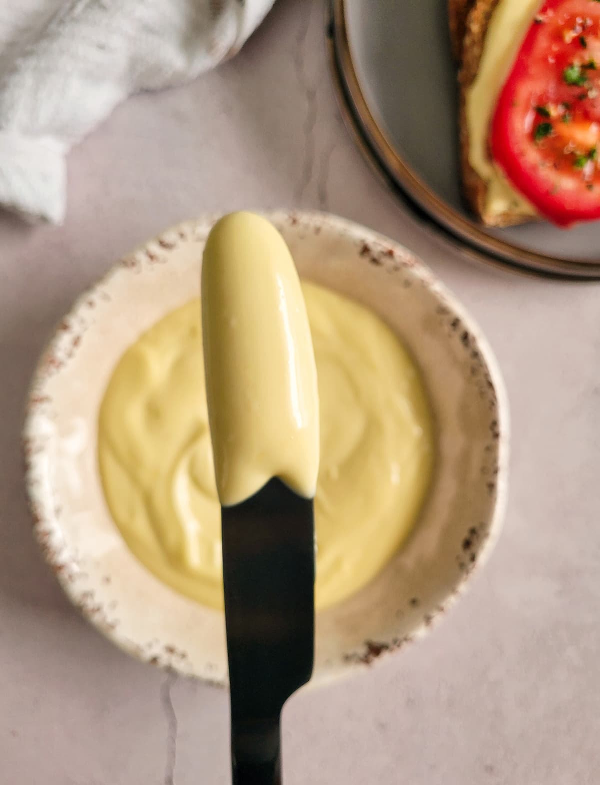 mayo on a black knife over a bowl of the rest of it, tomato on toast with mayo and parsley in the background