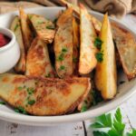 side view of a bowl of potato wedges garnished with fresh chopped parsley, ramekin of ketchup in the bowl, fresh sprig on parsley on the side