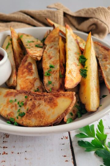 side view of a bowl of potato wedges garnished with fresh chopped parsley, ramekin of ketchup in the bowl, fresh sprig on parsley on the side