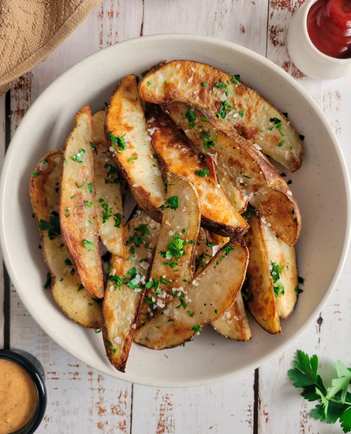 bowl of crispy baked potato wedges garnished with flaked salt and fresh chopped parsley, ketchup, bright orange dip and sprigs of fresh parsley in the background