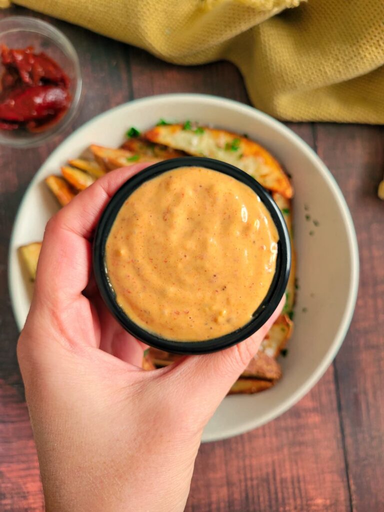 hand holding a ramekin of chipotle mayo over a bowl of potato wedges, ramekin of chipotle peppers in the background