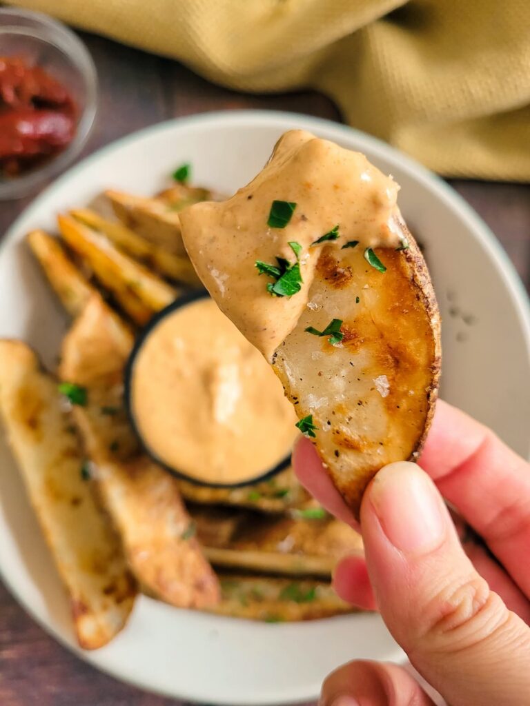 hand holding a half bitten potato wedge with orange sauce and fresh chopped parsley, more mayo, a bowl of wedges and chipotle peppers in the background