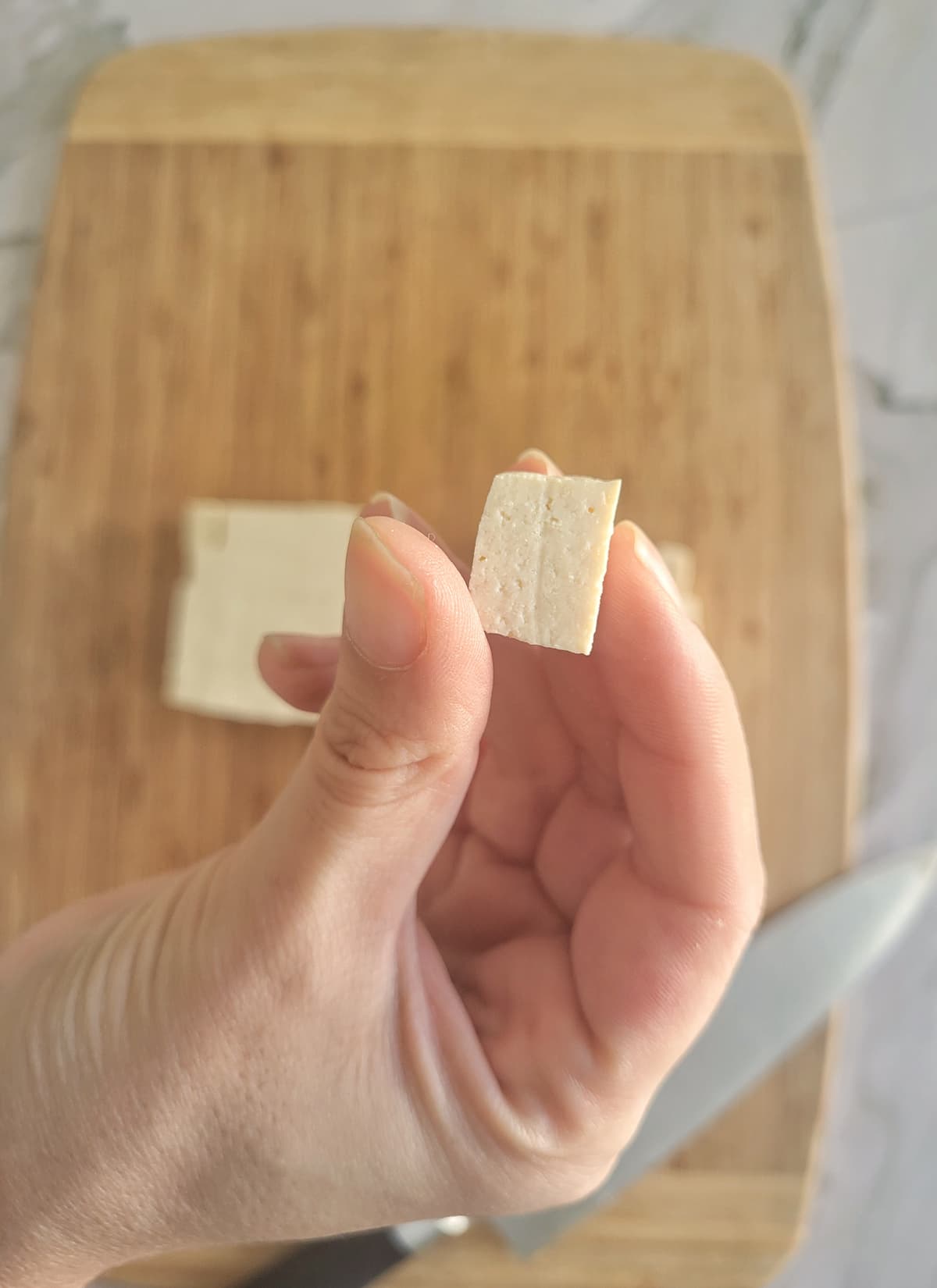 hand holding up a cube of tofu over a cutting board with a block of the rest of it