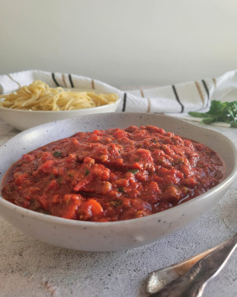 side view of a bowl of tomato sauce, fresh basil and bowl of pasta in the background