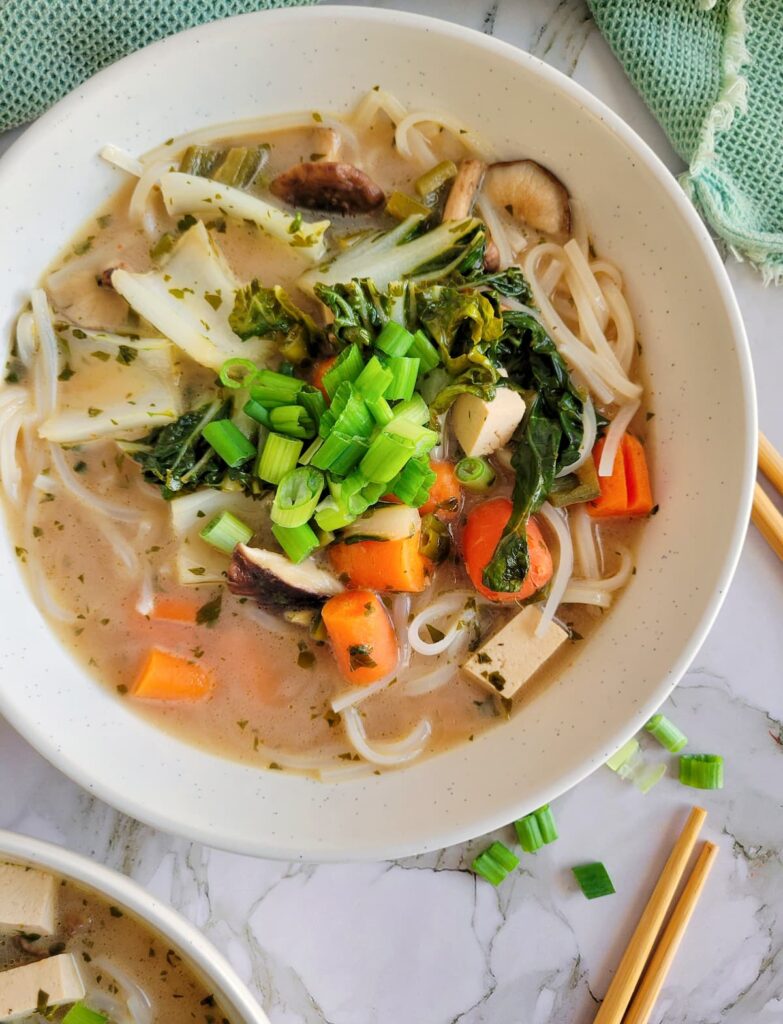 bowl of vibrant veggie soup in a creamy broth, chopped green onions, rice noodles, baby carrots, bok choy all in the bowl, chopsticks and chopped green onions in the background