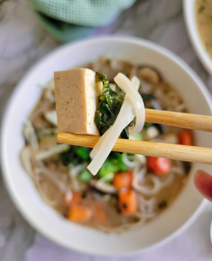 chopsticks holding up a piece of tofu, bok choy and rice noodle over a bowl of soup