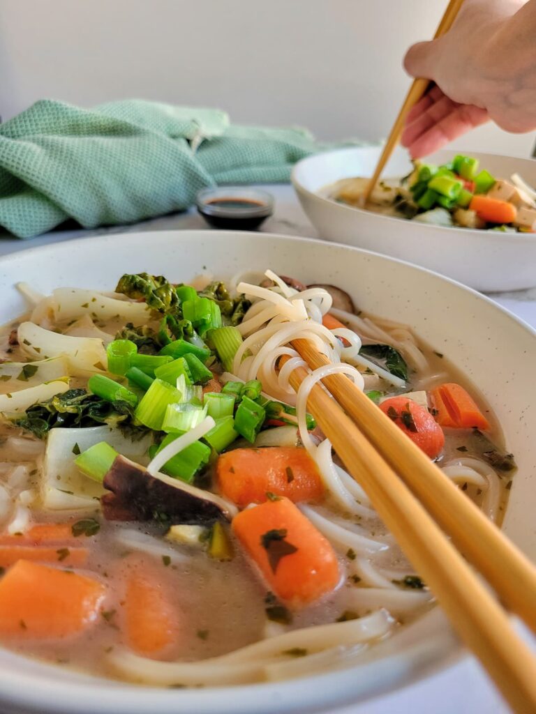 side view of veggie soup with baby carrots, green onions, mushrooms, another bowl of soup in the background with hands and chopsticks, soy sauce
