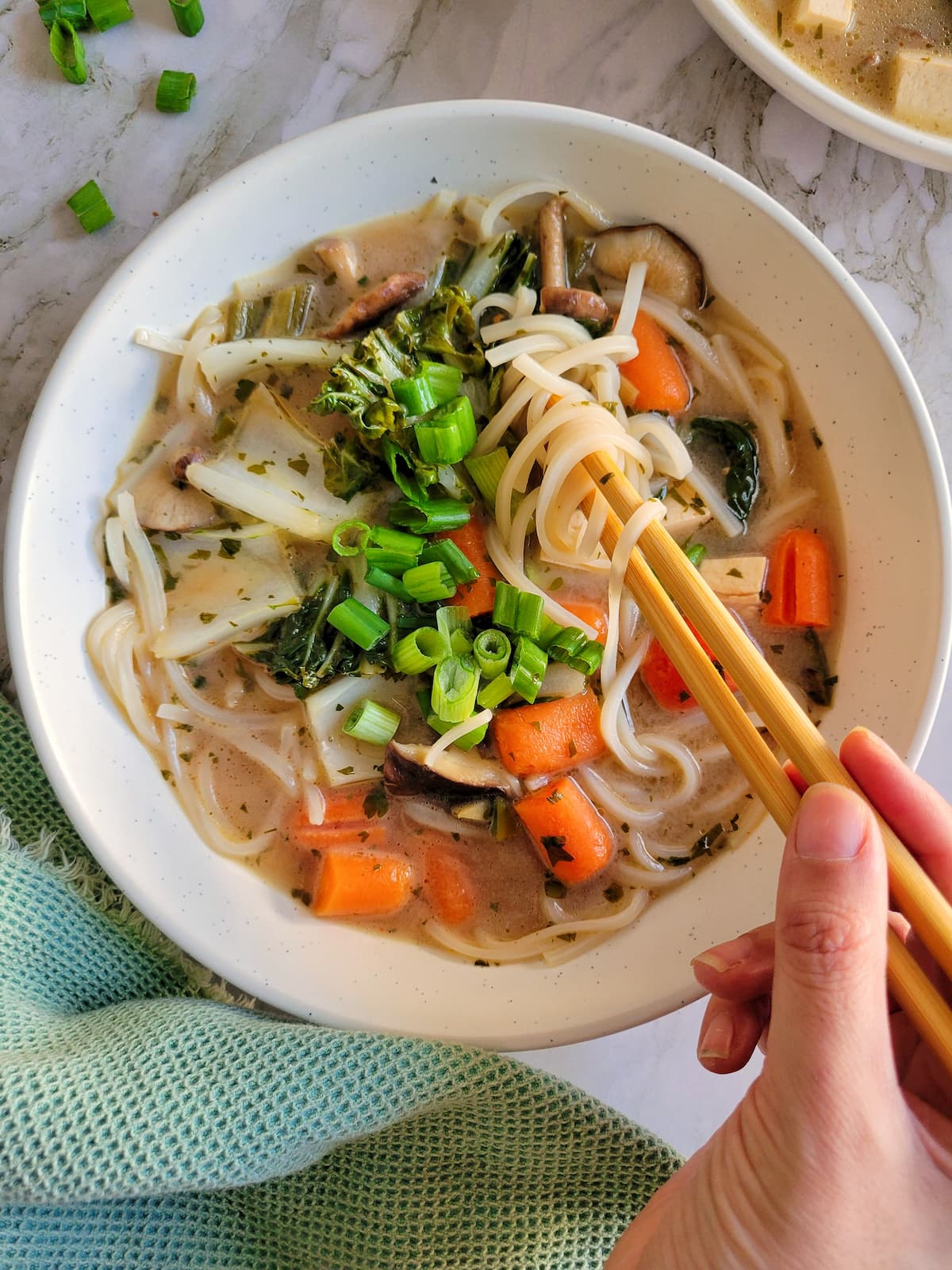 hand with chopsticks in a bowl of veggie soup with rice noodles, green onions, baby carrots, tofu and bok choy